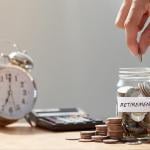 Hand putting coins in glass jar labeled "retirement" next to calculator and analog alarm clock with window in above in the background. 