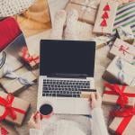 A shopper uses a laptop and credit card while sitting on a bed surrounded by Christmas gifts.