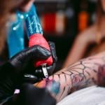 A professional artist tattooing a design on a woman's left forearm.