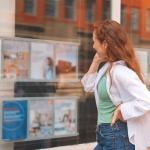 Woman with bright red hair explores property listings through a window while standing in front of a real estate display in a vibrant urban area.