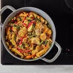 A colorful mix of stir-fried chicken, fresh vegetables, and cashews is sizzling in a pot on an induction cooktop. 