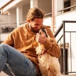 Sporty guy in honey-colored pullover hoodie and jeans sitting outside on steps playfully hugging a golden retriever.