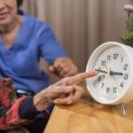 Close up of Asian elderly woman's hand pointing at analog clock with caregiver visible behind her.