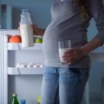Pregnant woman, whose face is not shown, is illuminated by light of open fridge holding a glass in one hand and bottle of milk in the other.