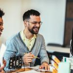 Group of three sitting at a table at work. Man in the middle is laughing and holding a pencil and two women are on either side of him.