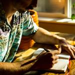 Man wearing a Hawaiian-style shirt sitting next to the window with light coming in, writing in a journal. 