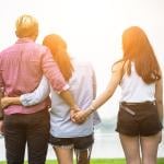 Couple with their back to the camera is looking at a scenic view with their arms around each other, but man is secretly holding hands with another person behind the woman's back.