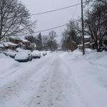 Unplowed snow accumulated on a residential street where rows of cars are parked.