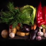 Assortment of beautifully-lit, fresh raw vegetables inlcuding leafy greens and red peppers in a wooden box against black background.