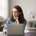 Woman sits at desk with open laptop computer, looking into the distance.