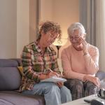 A senior woman and her caregiver sitting together on a sofa at home.