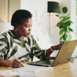 A black female bookkeeper working with a laptop and financial reports at home.