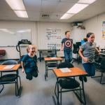 Three elementary-school-age students jumping in the air next to desks in a classroom. 