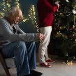 A stressed elderly man sitting by their Christmas tree as his partner decorates it.
