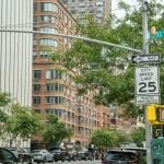 A City Speed Limit 25 sign with a photo enforcement notice stands near a traffic light on a busy urban corner. Modern buildings, cars, and a street sign for 53rd Street are visible in the background.