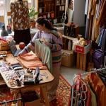 High-angle shot of sales assistant working at checkout desk cluttered with fashion accessories at second hand shop with retro style carpet selling clothes and household goods.