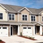 A row of modern townhomes with beige siding, white trim, and stone accents, each featuring a garage, driveway, and small landscaped front yards under a bright blue sky.