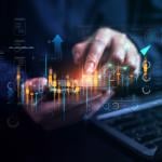 Stock market trading graphics illustrated over a businessperson using a phone and laptop.