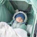 A baby in winter clothes sleeping in a green stroller outdoors.