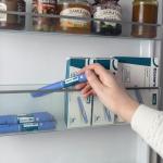 Close up of arm of person wearing white sweater reaching into refrigerator door to pick up an Ozempic injection pen, boxes of Ozempic are stored to the right.