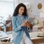 Fashion designer wearing powder-blue oversized blazer and white pants uses a tablet and tablet working logistics or small business managementwith office, shipping and clothing rack behind her.