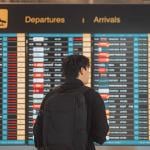 Rear view of a young man with a backpack looking at an airport's flight schedule board.