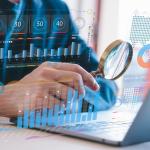 Person holding a magnifying glass close to a laptop screen, illustrated with data charts and graphics.