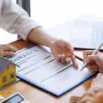Two people whose faces are not visible review paperwork, representing a real estate broker agent presenting form customer to sign for insurance agreement.