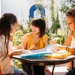 Three female university exchange students studying together around a sunny courtyard table.