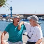 Two senior friends wearing caps and t-shirts in retirement life sitting on a bench at the harbor looking each other laughing happily.