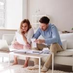 Two people sitting on a white sofa looking at bills and paperwork in a minimalistic living room.