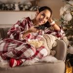 A brunette person enjoys popcorn during Christmas, lying in pajamas on the couch in front of the TV.