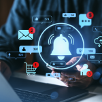 Person using a smartphone in front of a laptop with a various icons, like an alert bell, an email inbox notification, and a shopping cart, overlaying the image. 