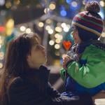 A mother kneels to the height of her child ourdoors and looks into his eyes, he is eating a heart-shaped lollipop and wearing a knitted cap with a pom pom in front of blurry christmas lights in the background.