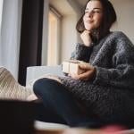 A young woman cozily sitting on a sofa at home holding a small gift box.