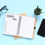 A pair of eyeglasses, a smartphone, and a notebook to list 2026 goals with a blue background.
