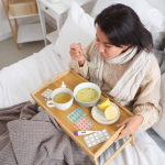 A woman sits in bed holding a spoon full of soup with a tray over her lap containing pills, nasal spray, a thermometer, lemons, a bowl of soup, and a mug of tea. 