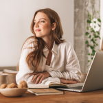 A content-looking woman in her 20s or 30s sits in a sunny room at a table with a laptop, a mug, a notebook, and a bowl of baked goods. 