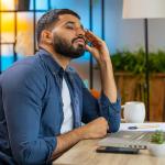 Man sitting at desk in modern colorful room looking at computer with an expression of distrust.