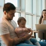 Woman smiling and sitting at table with laptop looking at man holding baby in the foreground.
