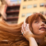 A woman's red hair flowing as she listens to her headphones outdoors.