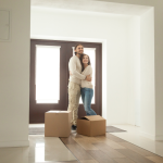A hugging couple stand in the house in front of the door with two cardboard boxes in front of them.