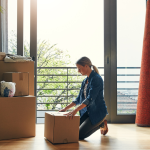 A women kneels in front of a cardboard box in a bright room with other boxes and household items like lamps and furniture. 