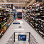 A shopping cart in a grocery store aisle filled with wine bottles.