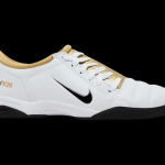 A pair of mostly white soccer shoes with a black Nike swoosh and a gold lining.