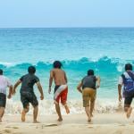 A group of five friends lined up to run towards a sea wave from the beach.