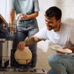 Happy man taking dishes out of the dishwasher while someone stands in the background.