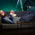 A bored man lying on couch and eating popcorn.