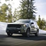 Low-angle view of earth-toned green 2026 Kita Sorento driving on mountain road with motion-blurred snow and pine trees in the background.