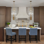A kitchen with wooden cabinets, modern gold fixtures and light blue velvet chairs around a center island.